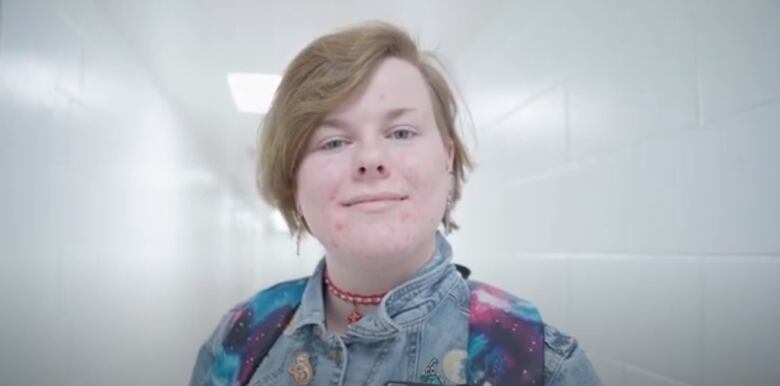 A woman with short blond hair, wearing a red necklace and a denim jacket, stands in a school hallway and looks to the camera.