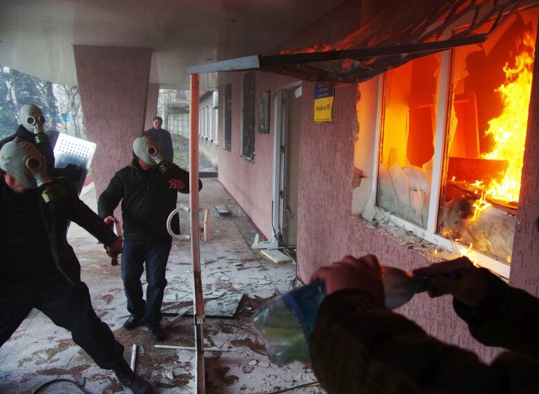 Protesters wear gas masks as one prepares a petrol bomb. The building next to them is on fire.