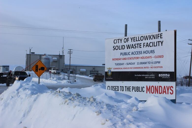 A sign on the right with information about Yellowknife's dump, surrounded by deep snow. A road, a truck and a building can be seen in the distance on the left. 