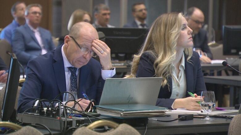 Man sits at a table intently staring at a laptop screen with a blonde woman beside him.