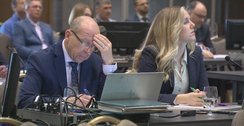 Man sits at a table intently staring at a laptop screen with a blonde woman beside him.