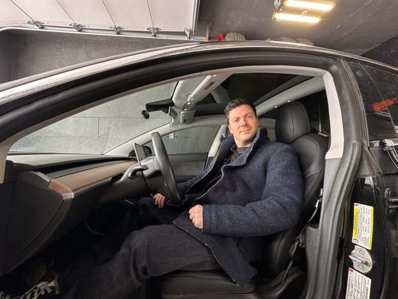 A white man in a dark blue trench coat sits in a car with the doop wide open. 