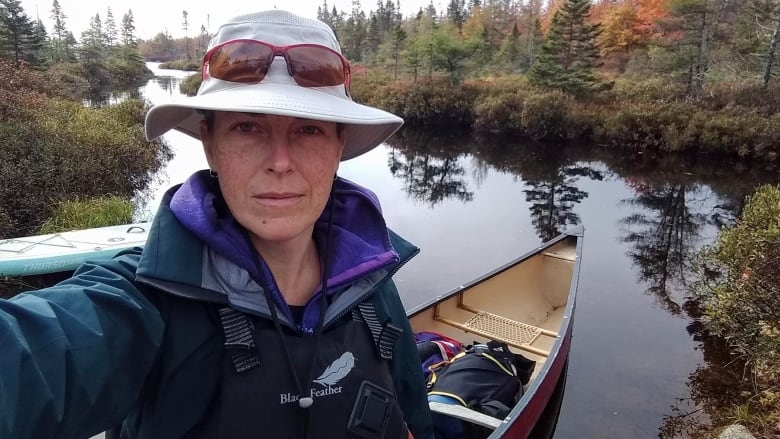 A woman takes a selfie. There's a canoe in the background, a body of water and trees.