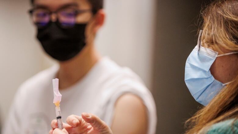 A person holds up a syringe beside a young person who is seated and wearing a mask. 