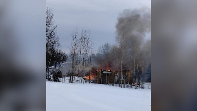 A structure is seen on fire, surrounded by snow, trees and dark grey smoke.