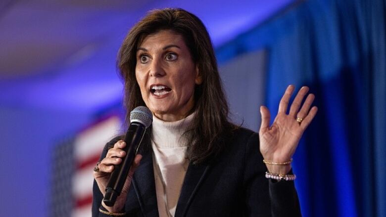 A woman wearing a brown blazer and beige turtle neck sweater speaks into a microphone.