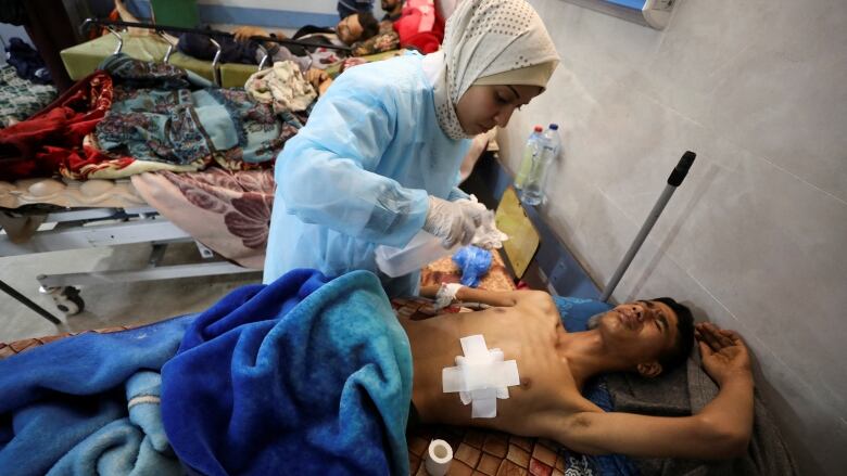 A health worker leans over a with a white bandage on his chest and expression of pain in a hospital bed.