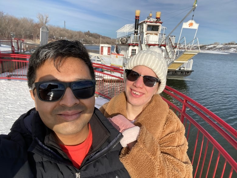 A man and a woman pose for a photo in front of a riverboat.