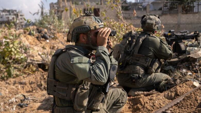 A soldier looks through binoculars as a second soldier holds a gun in the background.