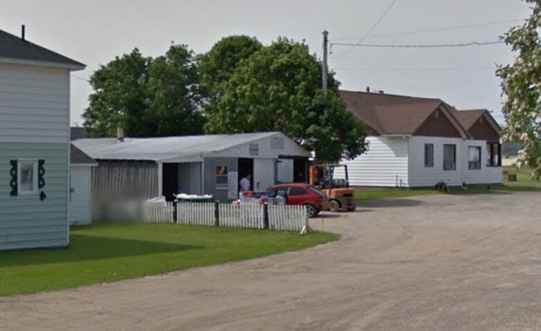 A person stands in the doorway of a one-story garage-type building with a small red car and a stack of boxes in front of it in a gravel driveway.