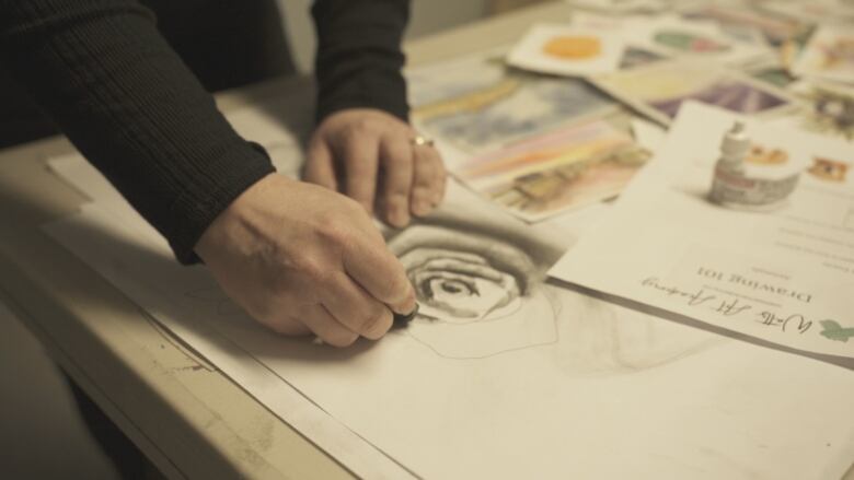 An artist with only her hands visible on screen is working on her sketch, which sits on a table full of sketches and stationary.  