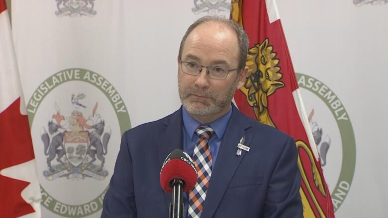 MacFarlane wears a plaid tie and blue shirt and coat. He stands against a backdrop of flags.