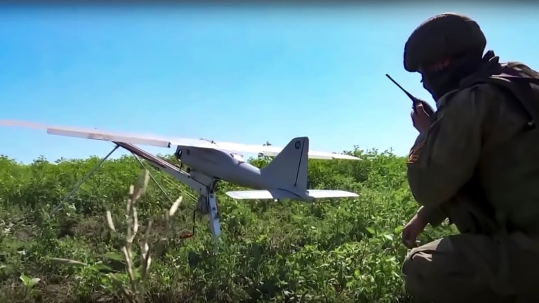 A soldier crouches behind a drone in a field.