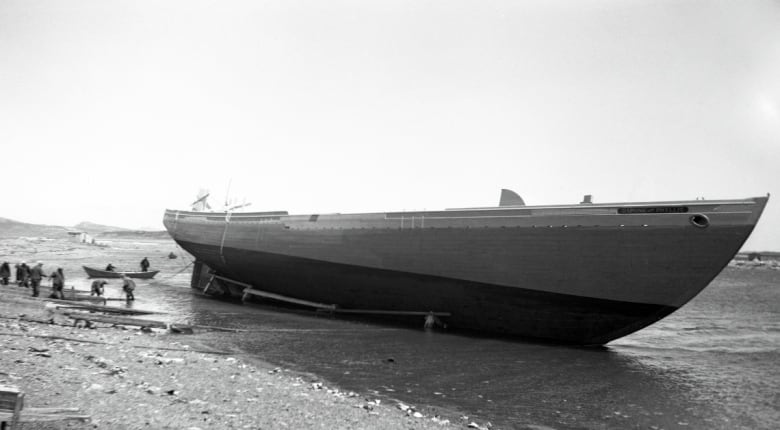 An old black and white photo showcases a large boat being launched into the water near a beach. A number of people are watching and assisting as the boat  slides further into the ocean. 