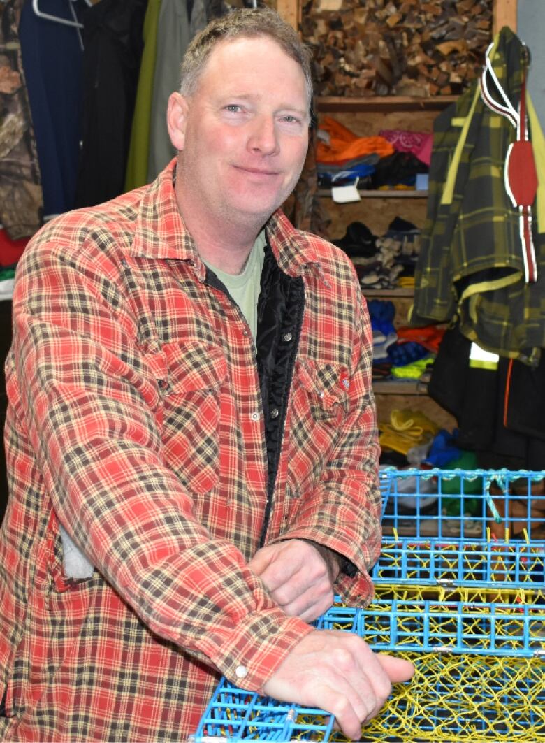 A middle aged man smiles as he rests his hands on a lobster pot that he is in the process of repairing.