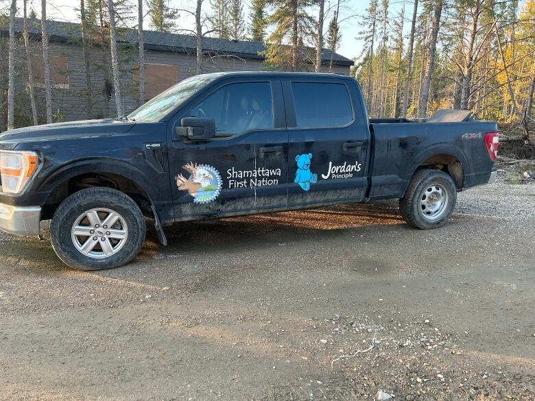 A black four-door pickup truck with emblems on the doors for Shamattawa First Nation and Jordan's Principle is parked with a row of trees and a building in the background.