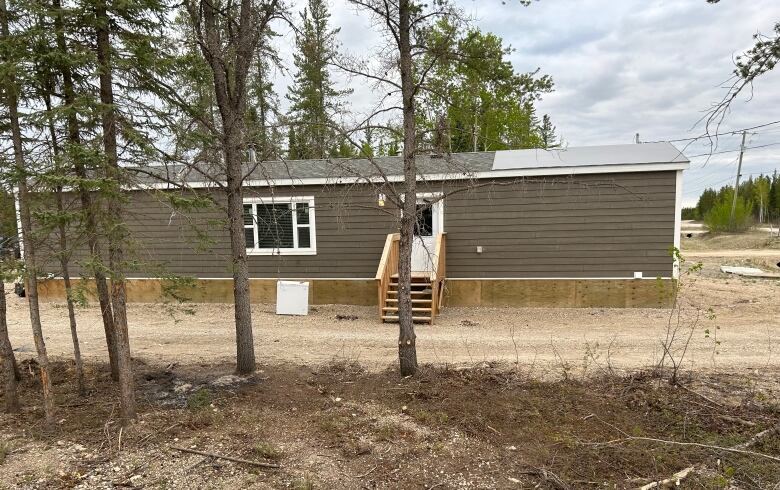 A taupe-coloured mobile home building has white trim around the window frame, a white door with stairs in front of it, and a row of trees in the foreground.