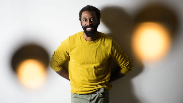 Ian Williams, a Black man, is standing in a mustard-yellow sweater with his hands behind his back. He has a beard, short hair and is standing between too blurry lights and a white background.