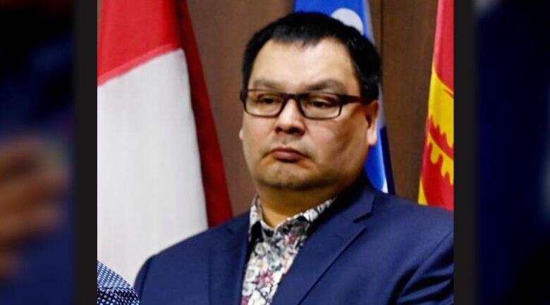 A man wearing a dark blue suit jacket and printed shirt is seen standing in front of flags in the background.