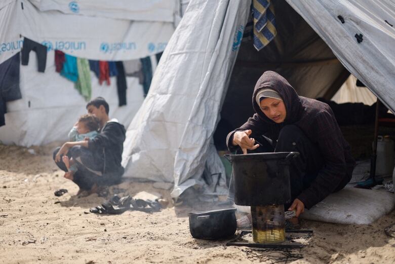 A woman cooks in a pot over a fire on the ground outside a tent.