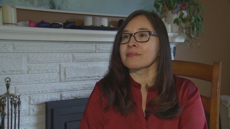 A woman wearing a red shirt and glasses sits next to a fireplace.