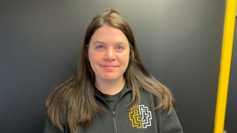 A woman with brown hair and a black half-zip sweater standing in front of a black wall. 