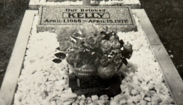 A photo of a grave to a dog named Kelly. It consists of a headstone and a bouquet of flowers over top of gravel