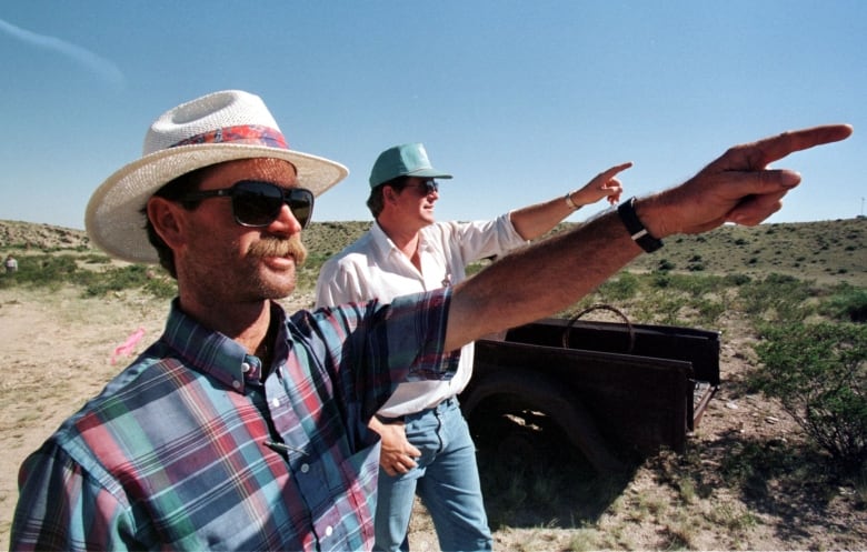 Two men in a desert point at  the sky