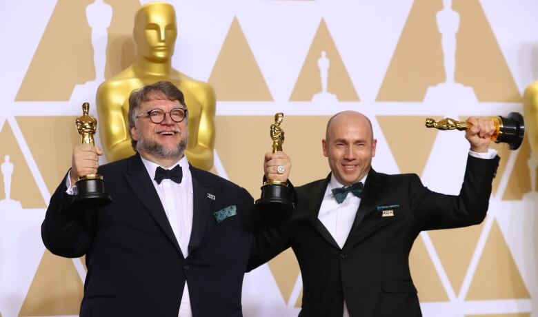 A bearded man with glasses and a bald man, both wearing tuxedos, hold up three Oscar statuettes in front of a larger ornamental Oscar statue. 