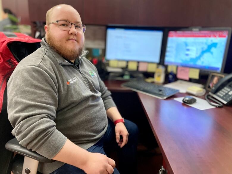 A bald man with a grey quarter-zip sweater sitting at a desk with computer screens in the background.