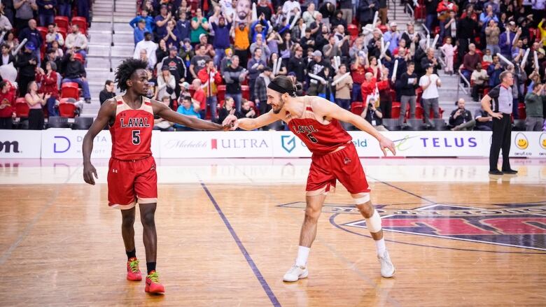 Two male basketball players shake hands in celebration on a court inside an arena filled with fans.