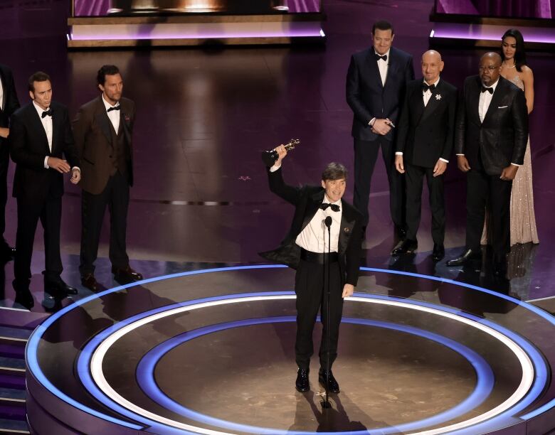 A man wearing a tuxedo raises an award in the air.