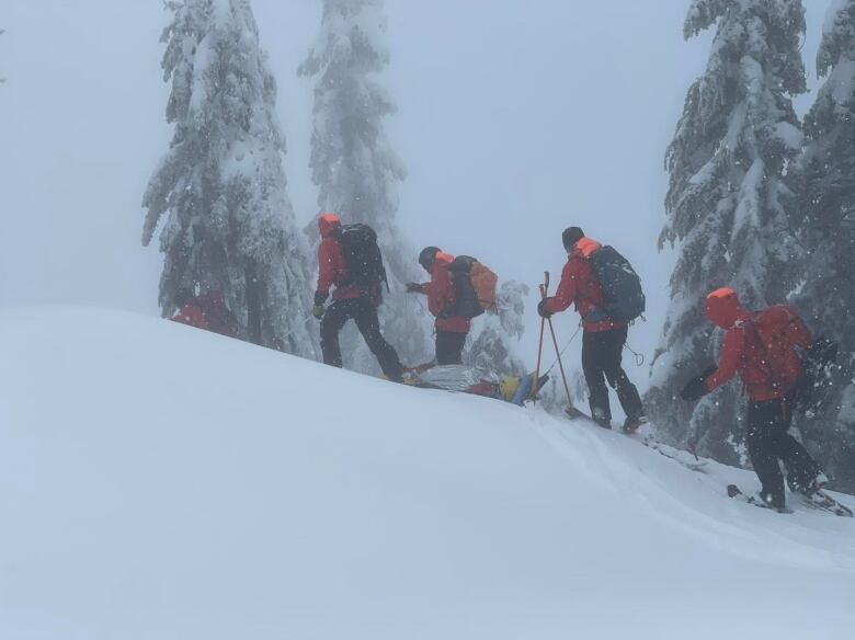 Rescuers wearing red jackets and backpacks are walking through the snow on skies and snowshoes. Between them is a woman wearing a helmet and covered in a foil-like material on a sled. 