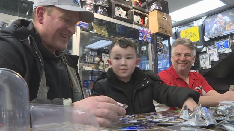 A young boy with his father and a store owner looking at a bunch of hockey cards stacked on a table. 