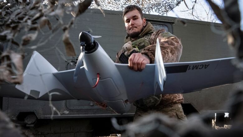 A soldier in camouflage uniform with a teddy bear tied to the front holds a drone that resembles a small airplane.