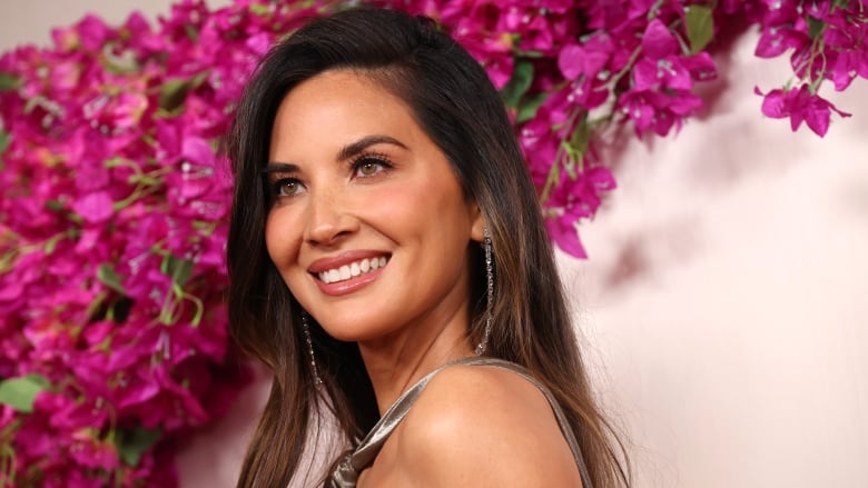 A smiling woman stands in front of a pink wall and flowers.