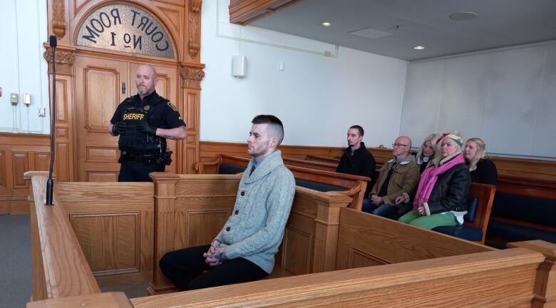 Man in pale sweater sitting in a box in a courtroom.