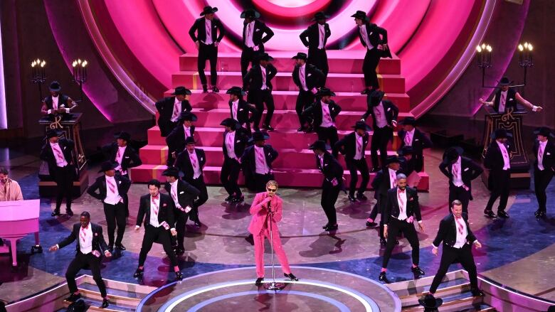 A stage full of men dressed in dark suits with pink sashes around a man singing into a microphone wearing a pink suit