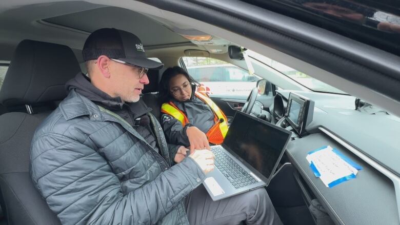 A man and a woman sit in the driver and passenger seat of a black SUV. The man has an open laptop. Both people are pointing at the laptop. 