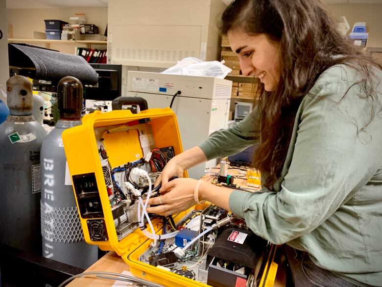 A woman in a laboratory works on a piece of scientific measurement equipment with hoses and fans encased in a hard plastic yellow case. 