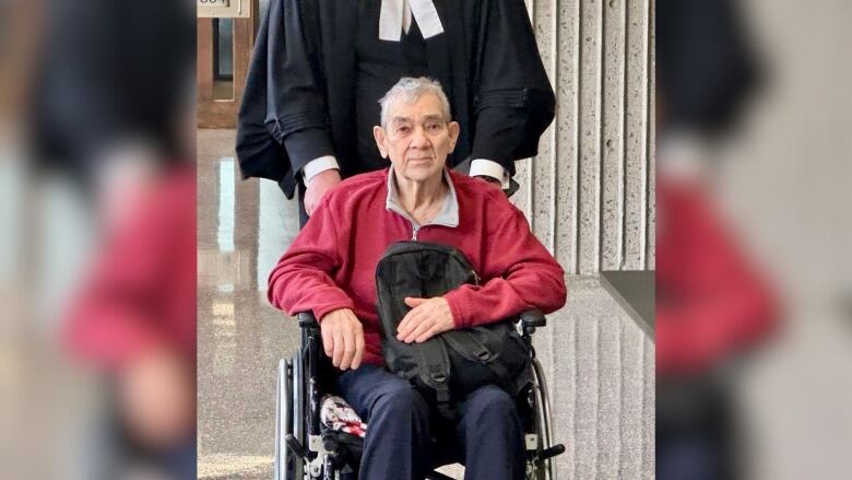 A man in a wheelchair is seen clutching a knapsack and being pushed by someone in a lawyer's gown.