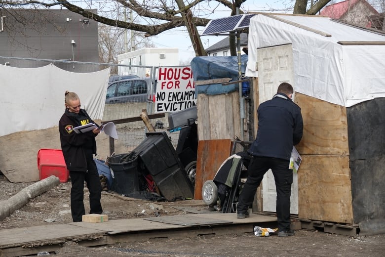 A female bylaw officer with a long blonde braid writes on a notepad, while a male officer attaches a bag with documents to the door of a small, white shack. A sign saying 