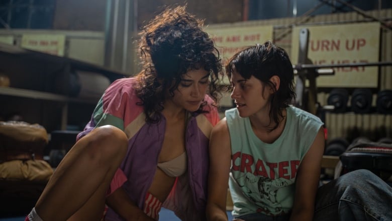 Two women wearing workout gear hold hands while sitting on the floor in a gym.