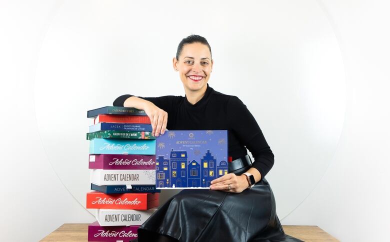 A woman smiling holding a box of chocolates while sitting on a bench next to a tower of chocolate boxes in front of a white background. 