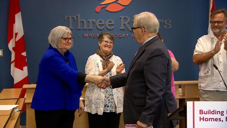 A woman and a man shake hands in front of a municipal sign while another woman smiles in the background. 