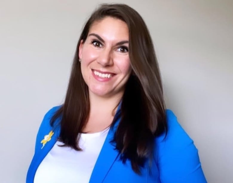 A woman in about her 30s with long brown hair and brown eyes smiles at the camera wearing a bright blue blazer with a yellow daffodil emblem on one lapel over a white t-shirt in an upper body portrait against a plain off-white background.