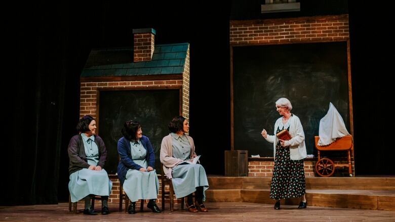 four actors performing on a stage. three women on the left are sitting and wearing the same outfit, a woman on the right is standing. 