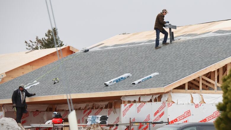 One man works at ground level on construction of a new house, while another man installs roofing materials on top.