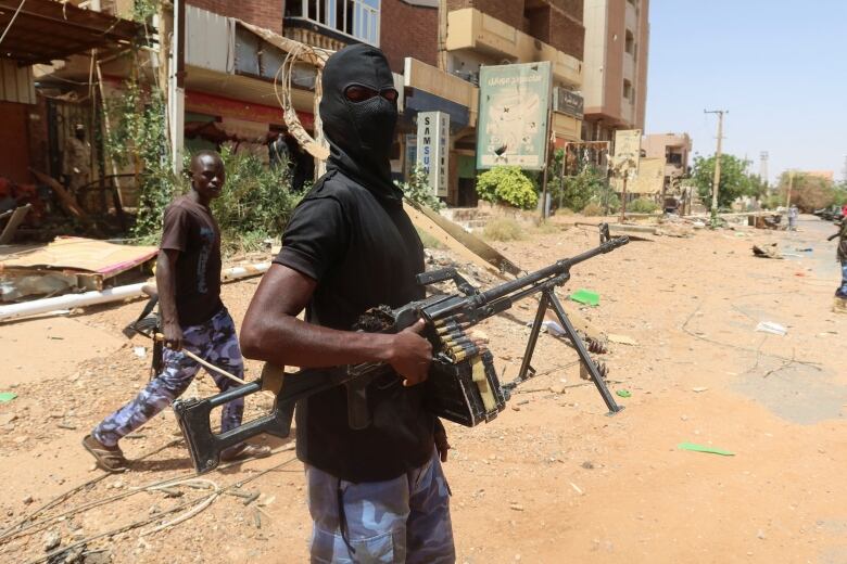 Two men walk down a garbage-strewn dirt road carrying large guns. Both are wearing blue camouflage pants and T-shirts and looking over their shoulders at the camera. One has a black face mask. 