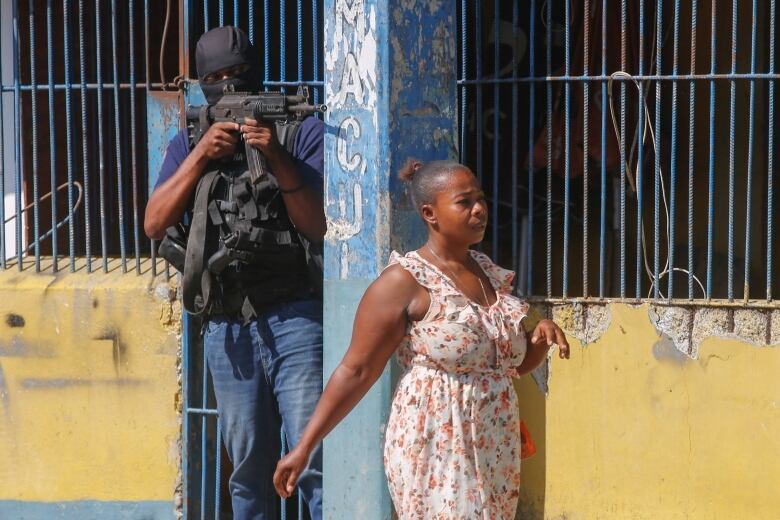 A woman walks past a masked police officer brandishing a rifle.